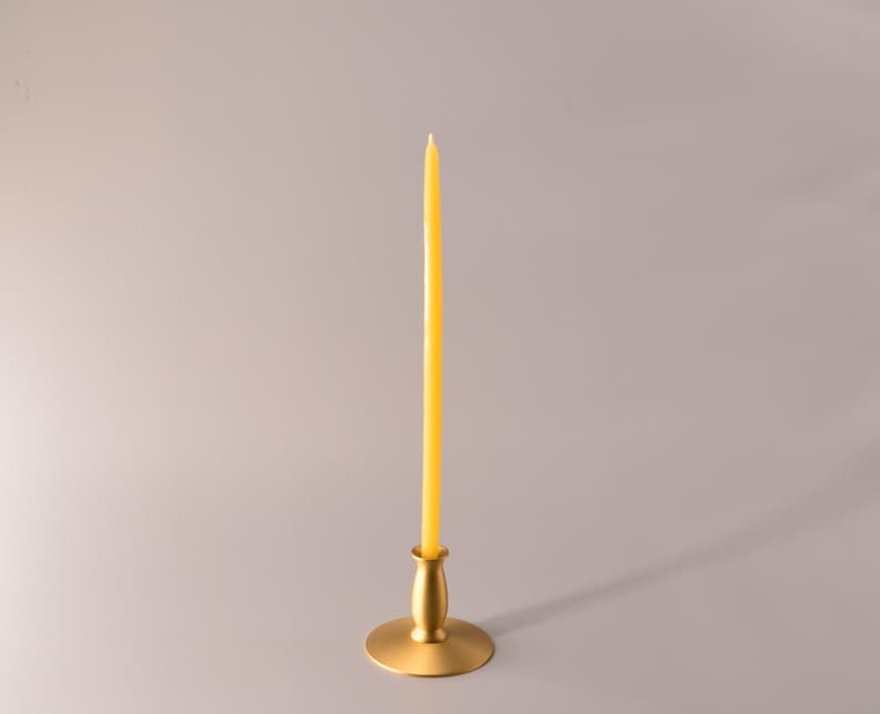 May include: A tall, slender yellow taper candle standing upright in a matte gold metal candlestick holder. The candle has a smooth, cylindrical shape with a pointed tip, and the holder features a wide, circular base and a tapered neck.