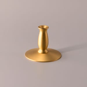 May include: A gold-colored metal candle holder with a wide, circular base and a central, tapered stem. The object features a smooth, matte finish and a small, cylindrical opening at the top designed to hold a single taper candle securely in place.