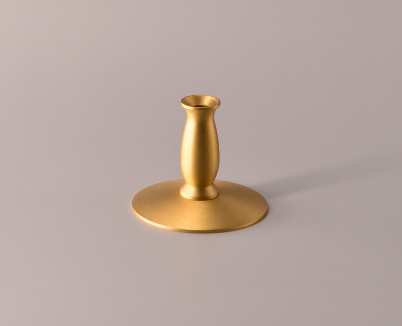 May include: A gold-colored metal candle holder with a wide, circular base and a central, tapered stem. The object features a smooth, matte finish and a small, cylindrical opening at the top designed to hold a single taper candle securely in place.