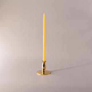 May include: A tall, slender yellow taper candle standing upright in a polished gold-tone metal candlestick holder. The candle is smooth and cylindrical, while the base of the holder is circular and flat, providing a stable foundation for the candle.