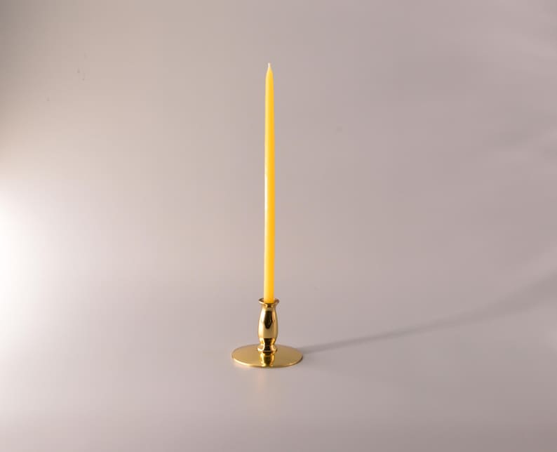 May include: A tall, slender yellow taper candle standing upright in a polished gold-tone metal candlestick holder. The candle is smooth and cylindrical, while the base of the holder is circular and flat, providing a stable foundation for the candle.