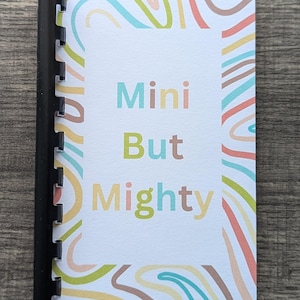 May include: A small, white notebook with a colorful abstract design on the cover. The notebook has a black spiral binding and the words "Mini But Mighty" printed in colorful letters on the front.