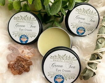 Könnte beinhalten: Drei kleine, runde, silberne Dosen mit grünem Tee-Augencreme. Jede Dose hat ein weißes Etikett mit den Worten "Beauty Fusion Green Tea Eye Cream" und "By- Jessica Hendershott". Jede Dose hat auch einen kleinen, runden, roten Aufkleber mit den Worten "Made in Montana USA".