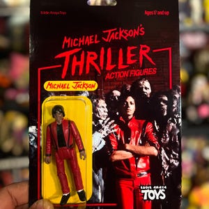 May include: A Michael Jackson Thriller action figure in a red jacket and pants, packaged on a black and red card. The card features the text "Michael Jackson's Thriller Action Figures" and "Eddie Anaya Toys."