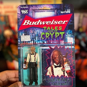 May include: A Budweiser Tales from the Crypt action figure in its original packaging. The figure wears a white shirt, black bowtie, and black trousers with suspenders, holding a miniature beer bottle. The card features the text Join me for a cold one.
