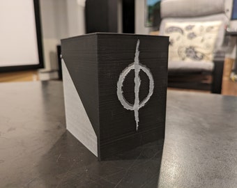 May include: A black cube with a white symbol on one side. The symbol is a circle with a vertical line through it.