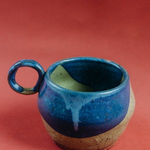 May include: A blue and brown ceramic mug with a unique, rounded handle. The mug has a speckled glaze and a light green interior.