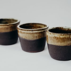 Puede incluir: Tres tazas de cerámica marrón y beige con un esmalte marrón oscuro. Las tazas están dispuestas en fila sobre una superficie blanca.