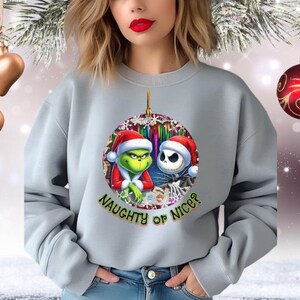 Puede incluir: Una sudadera gris claro con un dise&ntilde;o de adorno navide&ntilde;o con el Grinch y Jack Skellington con gorros de Pap&aacute; Noel. El adorno tiene el texto "NAUGHTY OR NICE?" en verde. La sudadera la lleva una persona.