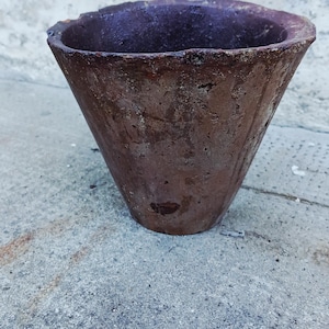 Op de afbeelding: Een conische plantenpot met een donkerbruin, verweerd en getextureerd oppervlak. De buitenkant vertoont tekenen van ouderdom met een ruwe, onregelmatige afwerking en een brede, open cirkelvormige rand. Het materiaal oogt dicht en solide.