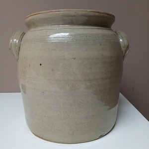 Può includere: Un grande vaso di ceramica di colore grigio chiaro con due manici. Il vaso ha una finitura smaltata ed è decorato con linee orizzontali.