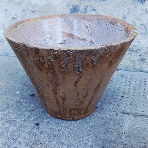 May include: A conical terracotta bowl with a weathered, rustic finish. The exterior features a textured, mottled brown surface with visible drip-like patterns, while the interior has a lighter, worn, and uneven off-white glazed appearance.
