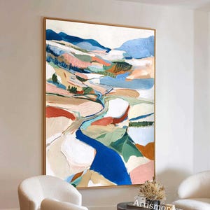Puede incluir: Pintura abstracta de paisaje con un río de pintura azul que fluye a través de un valle de tonos rosas, verdes y marrones. La obra de arte está enmarcada en un marco dorado y cuelga de una pared blanca. La pintura se exhibe en un salón con sillas blancas y una mesa de madera.