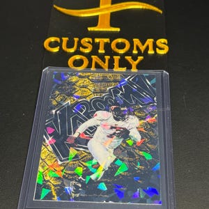 May include: A collectible sports trading card encased in a clear plastic protective sleeve. The card features a football player in a white uniform against a gold and black background with the word KABOOM in large letters and holographic foil accents.