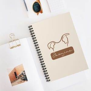 May include: Cream-colored spiral notebook with a brown line drawing of a dog and the name "Jasmine" in script. The notebook is open, revealing a blank page. A gold binder clip and sunglasses are also visible.