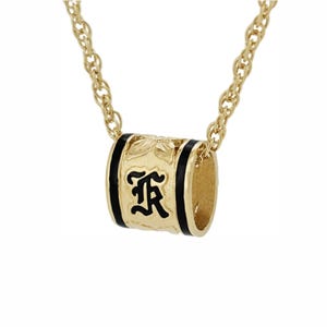 May include: Gold-tone necklace featuring a cylindrical pendant with black enamel accents. The pendant has a stylized letter "R" in black and floral detailing. The necklace chain is a delicate rope style.