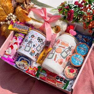 May include: A pink gift box containing a selection of treats, including two white tumblers with food-themed designs, sweets, and coffee pods. The box also holds a white plush item with a pink ribbon and a gold spoon.
