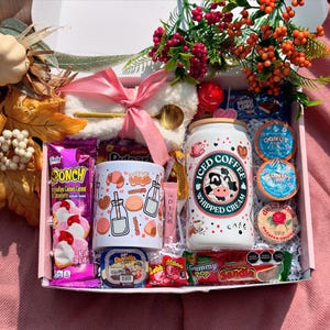 May include: A gift box containing a mug with pastry illustrations, a can of iced coffee with a cow design, and assorted sweets. The box is adorned with a pink bow and floral decorations, presenting a celebratory arrangement.