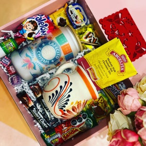 May include: A pink gift box containing Mexican snacks and treats. It features a mug with floral designs and the words "Cafecito y Chisme", a jar, sweets, chocolate, and a packet of Abuelita hot chocolate mix.