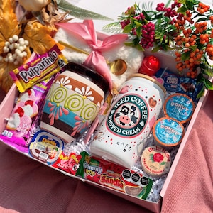 May include: A gift box containing a decorated ceramic mug, a can of iced coffee, sweets, and donut coffee pods. The box is adorned with a pink ribbon and flowers. The mug is decorated with a floral design.