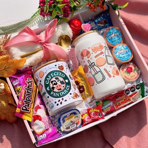 May include: A gift box containing a variety of treats, including two white tumblers with food-themed designs, sweets, and coffee pods. The box also features a plush toy, a lollipop, and decorative flowers. The tumblers have bamboo lids.