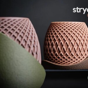 May include: Two decorative planters with a geometric lattice design. One is a dusty rose colour, and the other is a terracotta colour. The planters are labelled "stryde planter" in white text. The planters are on a dark surface.