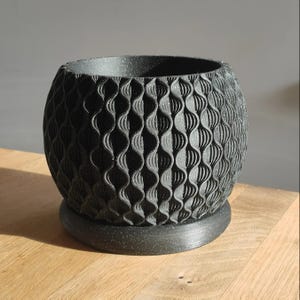 May include: A black, spherical planter with a textured, wave-like pattern. The planter sits on a matching round base. The design is modern and geometric, suitable for indoor or outdoor use. The planter is made of a speckled material.