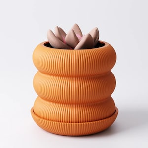 May include: A terracotta-coloured, ribbed ceramic plant pot with a matching saucer. The pot has a distinctive, stacked design and holds a faux succulent with pink and beige petals. A modern piece for home decor.