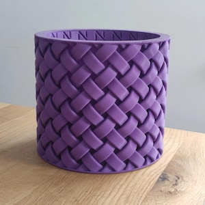 Pode incluir: Um vaso cilíndrico roxo com um design de cesta tecida. O vaso é feito de um material sólido e tem uma textura suave. A borda superior tem um padrão decorativo. Este vaso decorativo é adequado para uso interno ou externo.