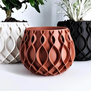 Pode incluir: Um vaso de cor terracota com um padrão geométrico repetitivo é o ponto focal. Ao lado, há vasos brancos e pretos, cada um com vegetação. Os vasos estão sobre uma superfície branca, criando uma estética limpa e moderna.