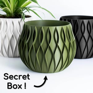 May include: Three decorative plant pots in white, olive green, and black. The olive green pot is in the foreground, with a textured, wave-like pattern. The text "Secret Box!" is visible. The pots are designed for indoor or outdoor use.