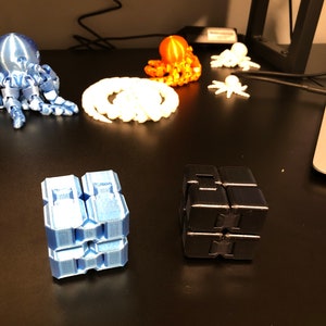 Op de afbeelding: Een blauwe, zwarte en oranje 3D-geprinte octopus, een witte 3D-geprinte octopus en twee 3D-geprinte fidget cubes, een blauwe en een zwarte.