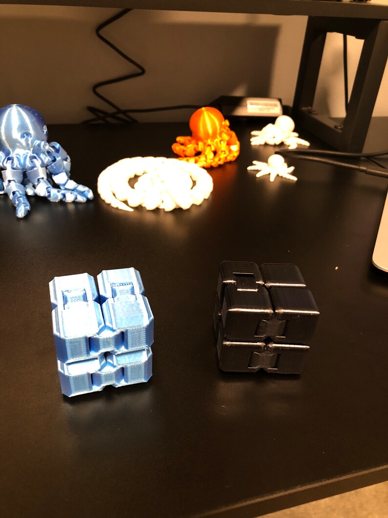 Op de afbeelding: Een blauwe, zwarte en oranje 3D-geprinte octopus, een witte 3D-geprinte octopus en twee 3D-geprinte fidget cubes, een blauwe en een zwarte.