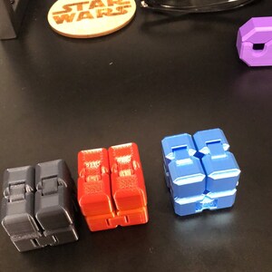 Op de afbeelding: Vier fidget cubes in verschillende kleuren: zwart, rood, blauw en paars. De kubussen zijn gemaakt van plastic en hebben een getextureerd oppervlak.