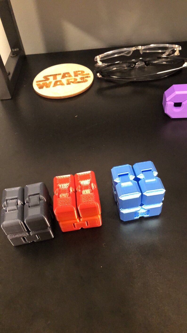 Op de afbeelding: Vier fidget cubes in verschillende kleuren: zwart, rood, blauw en paars. De kubussen zijn gemaakt van plastic en hebben een getextureerd oppervlak.