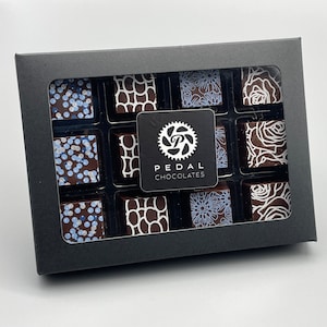 May include: A black box with a clear window showing nine assorted chocolate squares. The chocolates are decorated with white designs, including flowers, geometric patterns, and sprinkles. The box has a white label with the text "PEDAL CHOCOLATES" and a bicycle gear logo.