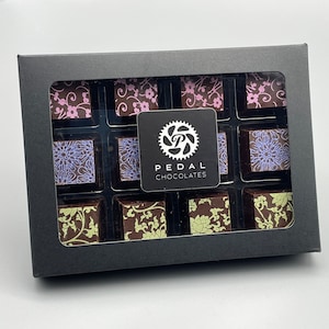 May include: A black box with a clear lid containing 12 assorted chocolate squares. The chocolates are decorated with floral patterns in shades of pink, purple, and green. The box has a white label with the text "PEDAL CHOCOLATES" and a gear logo.