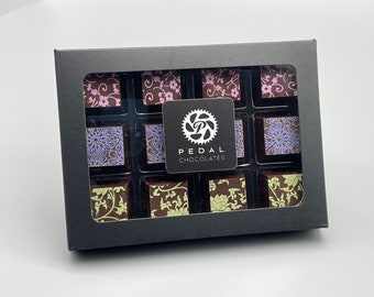 May include: A black box with a clear lid containing 12 assorted chocolate squares. The chocolates are decorated with floral patterns in shades of pink, purple, and green. The box has a white label with the text "PEDAL CHOCOLATES" and a gear logo.