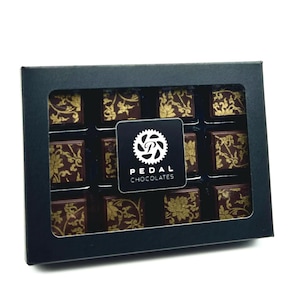 May include: A black box with a clear window showing 12 chocolate squares. Each square has a gold design and the box has a logo that says "PEDAL CHOCOLATES".