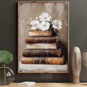 May include: An oil painting depicts a stack of antique books topped with white flowers. The books are brown and beige, with visible text on the spines. The artwork is framed in a wooden frame and has a neutral color palette.