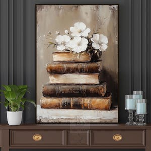 May include: An oil painting depicts a stack of antique books topped with white flowers. The books are various shades of brown and tan, with visible text on the spines. The artwork is framed in a dark brown frame.