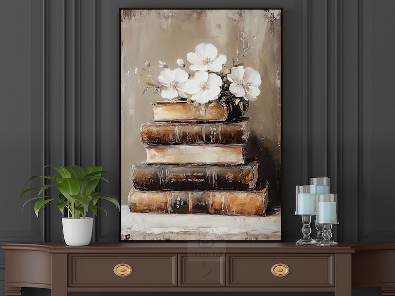 May include: An oil painting depicts a stack of antique books topped with white flowers. The books are various shades of brown and tan, with visible text on the spines. The artwork is framed in a dark brown frame.