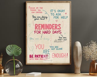 May include: A framed print with inspirational text in pink and teal. The words "REMINDERS FOR HARD DAYS" are prominent, with phrases like "You are doing your best" and "You are enough!" The artwork is displayed on a wooden table.