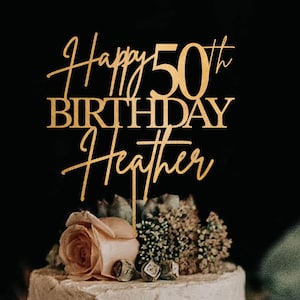 May include: A gold-colored cake topper that reads "Happy 50th Birthday Heather" in elegant script. The topper is placed on a white frosted cake decorated with flowers and greenery, set against a dark background.