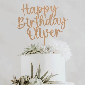 May include: A wooden cake topper with the text "Happy Birthday Oliver" in a cursive font. The topper is brown and has a rustic look. It is placed on top of a white cake decorated with succulents.