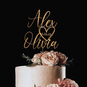 May include: Gold wedding cake topper with the names Alex and Olivia and the date 2.25.23 inside a heart.