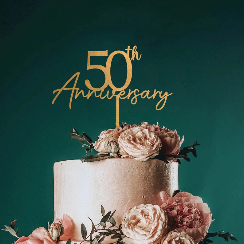 May include: Gold 50th Anniversary cake topper with the words "50th Anniversary" in a cursive font. The topper is placed on a two-tiered white cake decorated with pink roses and greenery.