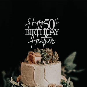 Pu&ograve; includere: Una torta di compleanno con un cake topper che recita "Happy 50th Birthday Heather". La torta &egrave; decorata con fiori e verde. La torta ha una glassa di colore chiaro. Lo sfondo &egrave; scuro.