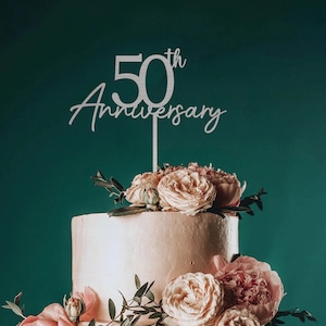 May include: A white cake topper with the text "50th Anniversary" in a cursive font. The topper is placed on a two-tiered white cake decorated with pink roses and greenery.