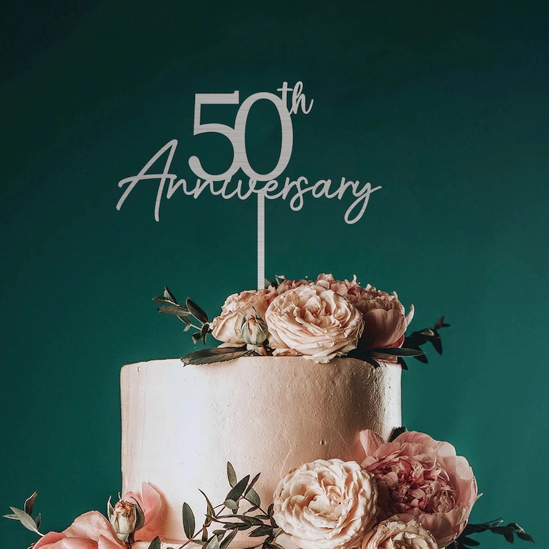 May include: A white cake topper with the text "50th Anniversary" in a cursive font. The topper is placed on a two-tiered white cake decorated with pink roses and greenery.